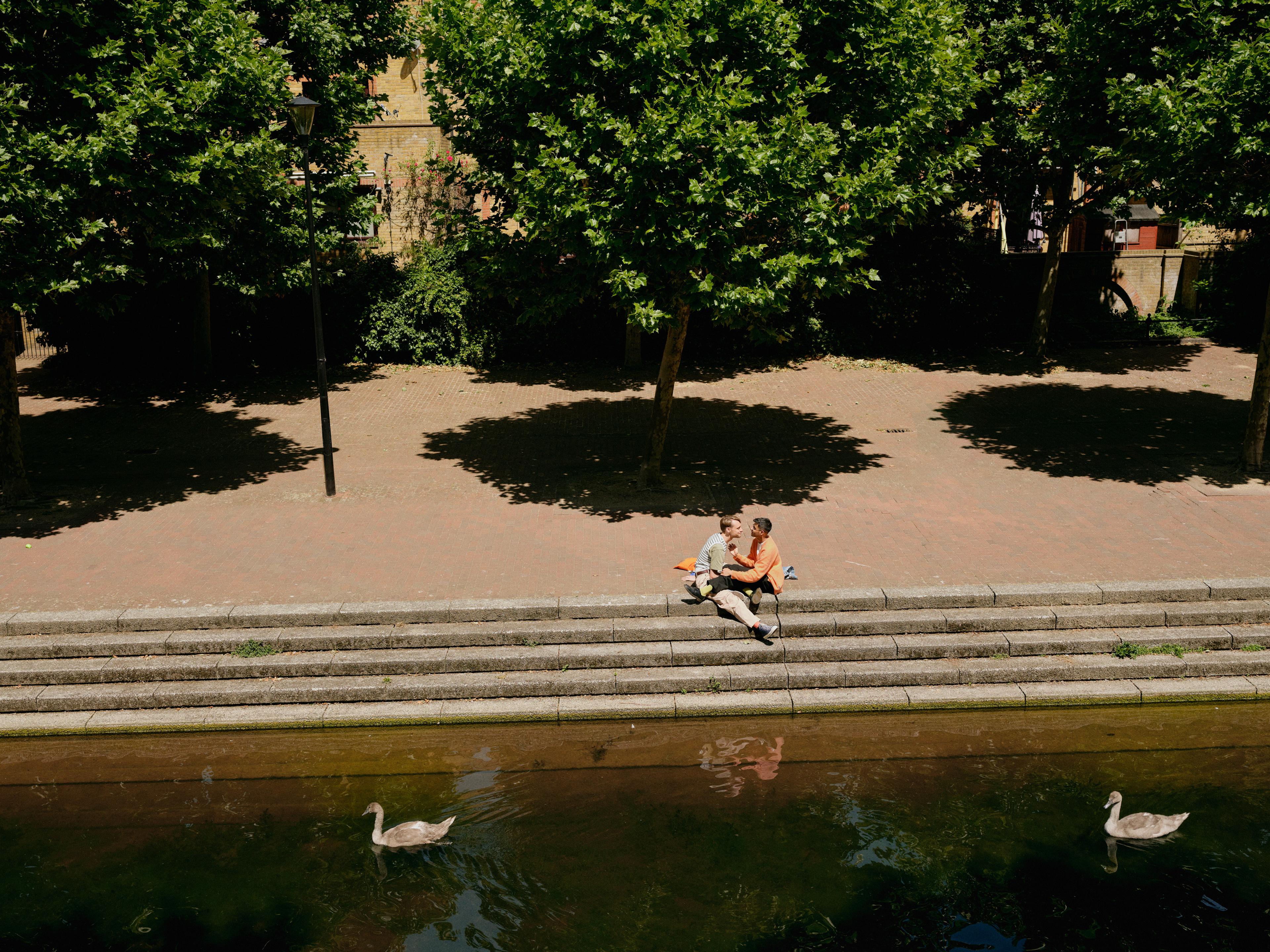 Two people on a date on top of some steps next to a body of water, there are two ducks swimming by and trees behind them.