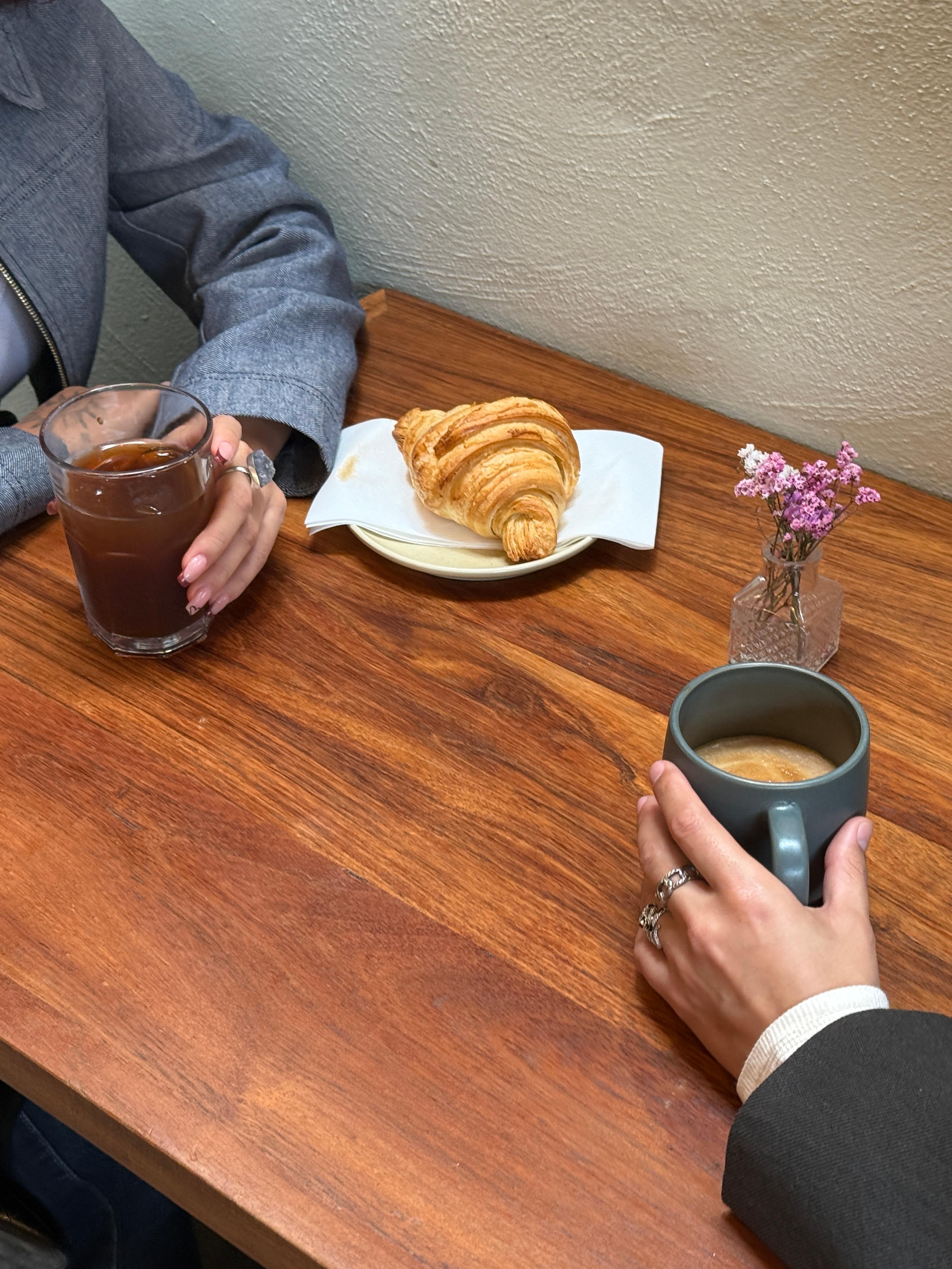 Two people sitting across from each other at a table, each holding a beverage. On top of the table is a croissant and some flowers.