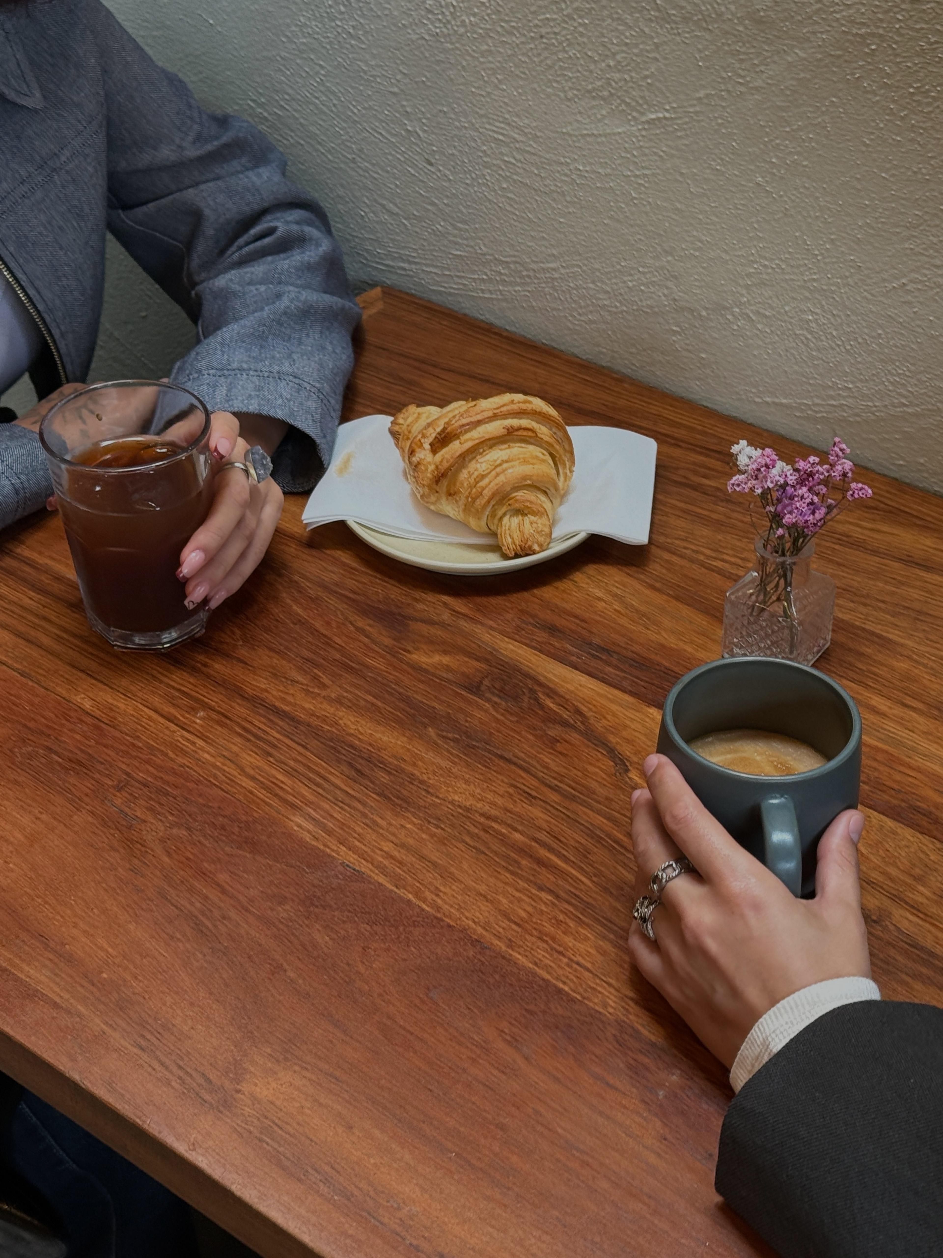 Two people sitting across from each other at a table, each holding a beverage. On top of the table is a croissant and some flowers.
