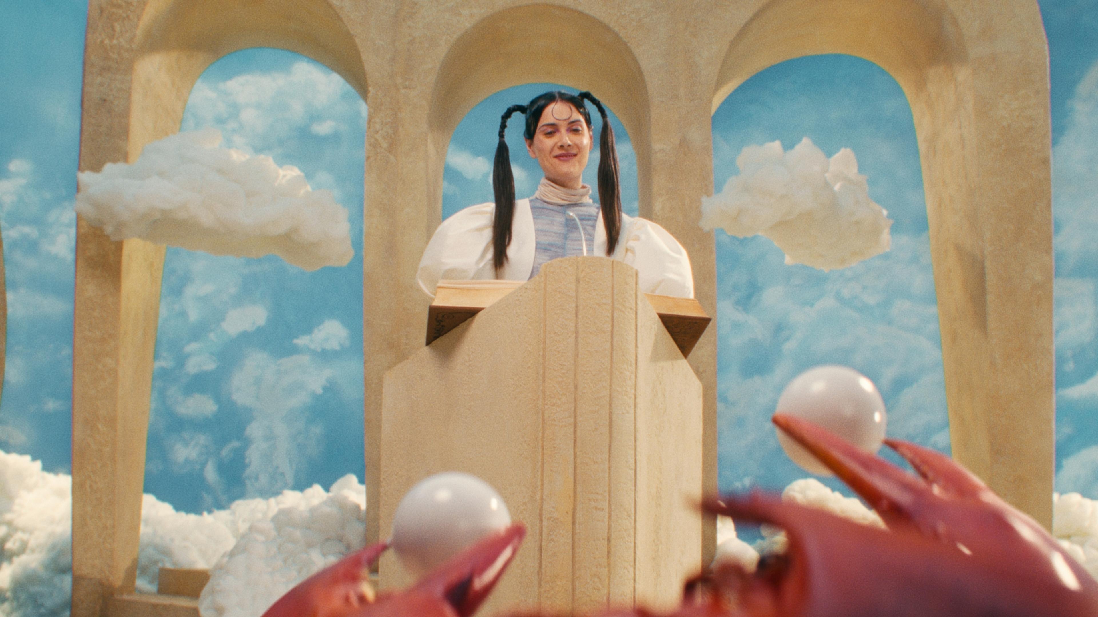 A person in pigtails stands behind a podium bearing a book, arches and clouds are visible behind them.