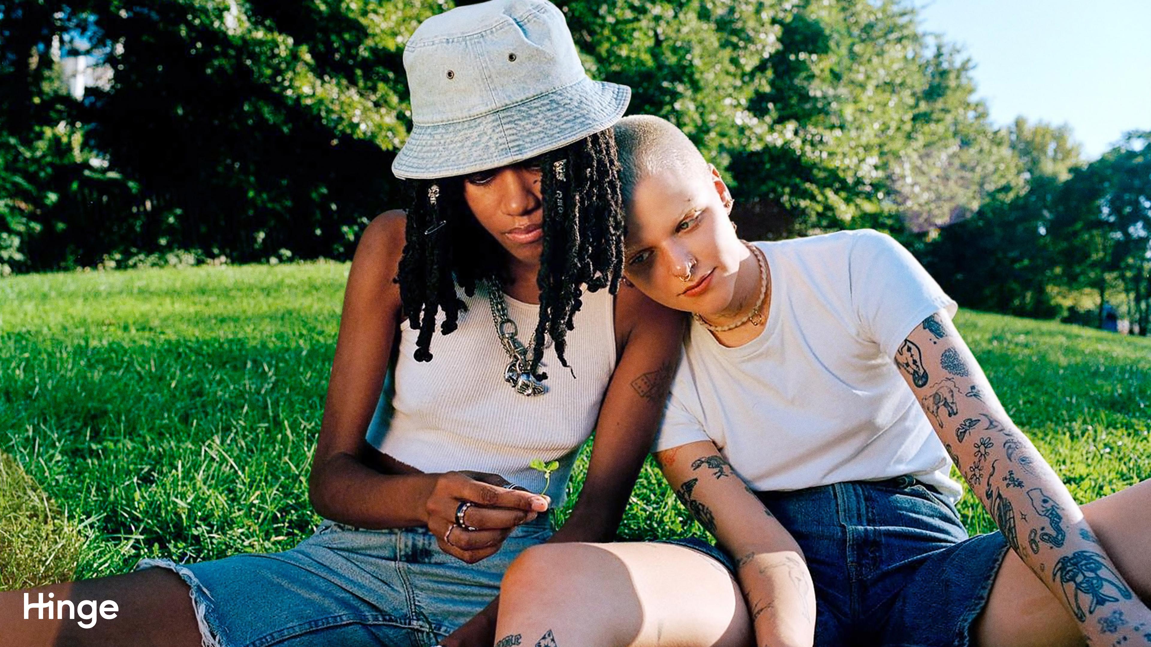 Two people sit in a field of grass, one leans on the others shoulder.